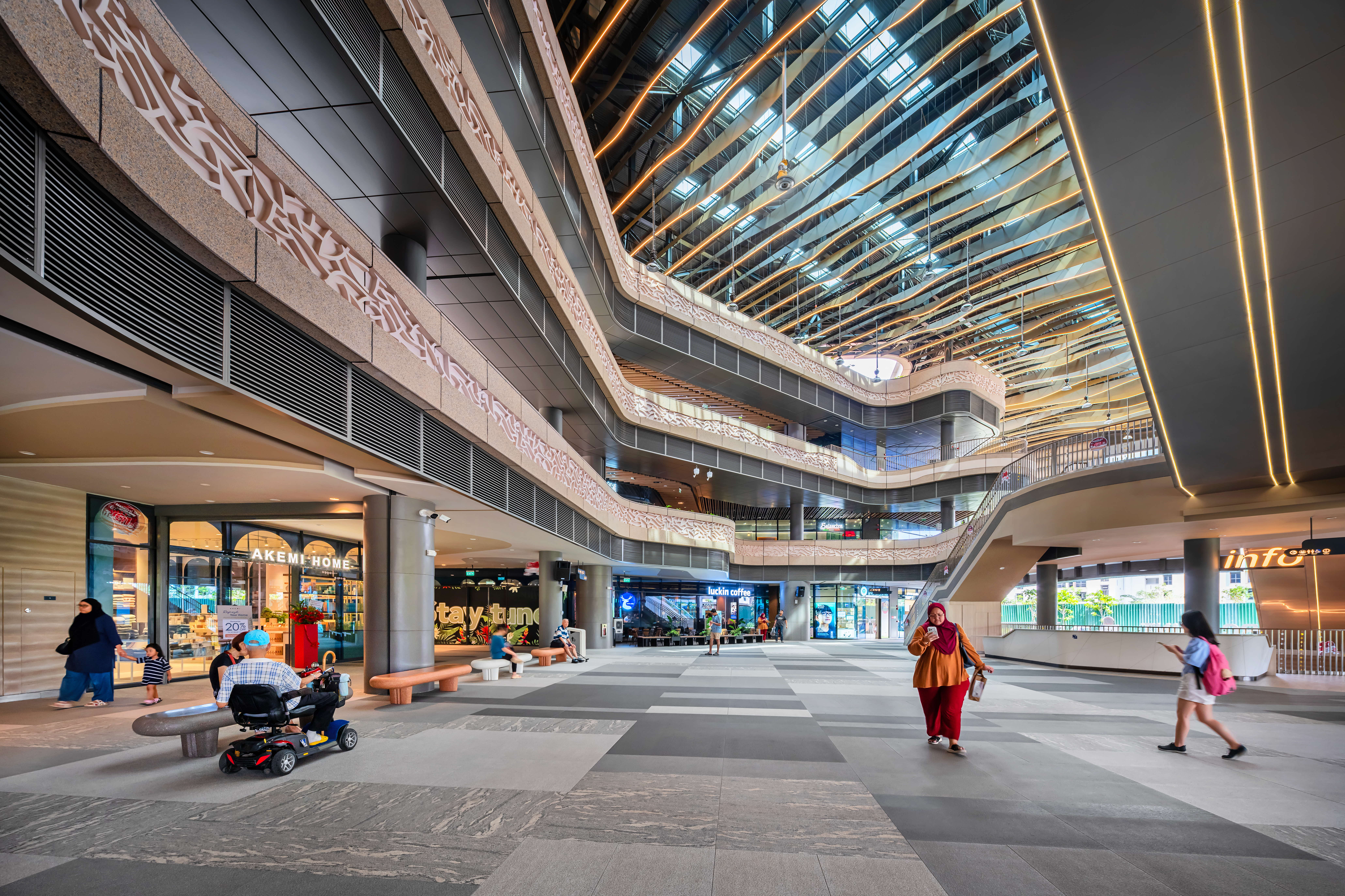 Open atrium mall with glass ceiling, multiple floors, escalators, shopfronts, and people walking and sitting